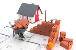 Modell von einem Einfamilienhaus, Ziegeln und Schubkarre auf einem Bauplan.